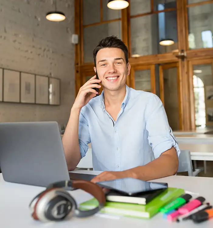 A smiling young man in a light blue shirt talking on his phone at a modern workspace with a laptop, headphones, and colorful highlighters on the table in front of him