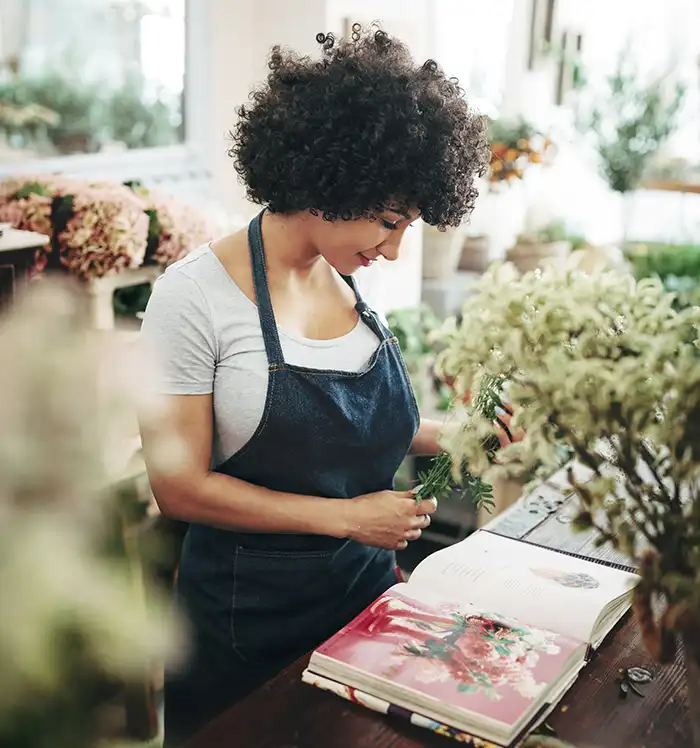 A smiling woman with curly hair wearing a denim apron holding an open book in a charming plant-filled shop, laughing with her head tilted back