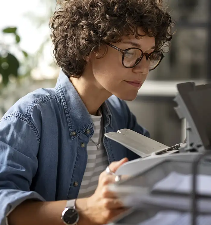 A confident woman with curly hair and glasses sitting at a wooden desk in a bright modern office, looking thoughtfully to the side with a printer visible in the background
