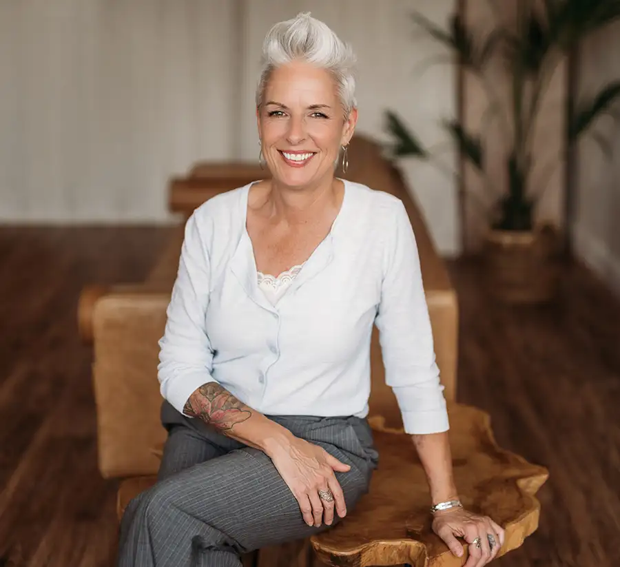 Nadine Hill, Dover NH bookkeeper and founder of Balance in Your Business, smiling warmly while seated on a rustic live-edge wooden bench, wearing a soft white cardigan and grey trousers, with short silver hair, sleeve tattoo visible, in a bright airy room with warm wood floors and a large plant in the background