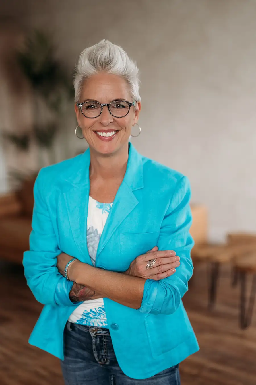 Nadine Hill, Dover NH bookkeeper and founder of Balance in Your Business, smiling confidently with arms crossed, wearing a bright teal blazer and glasses, with short silver hair, standing in a warm, softly lit room