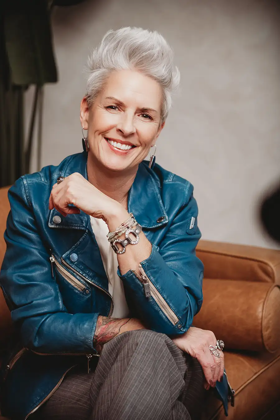 Nadine Hill, Dover NH bookkeeper and founder of Balance in Your Business, smiling warmly while seated on a caramel leather sofa, wearing a blue leather jacket and statement jewelry, with short silver hair, sleeve tattoo visible, in a softly lit studio setting