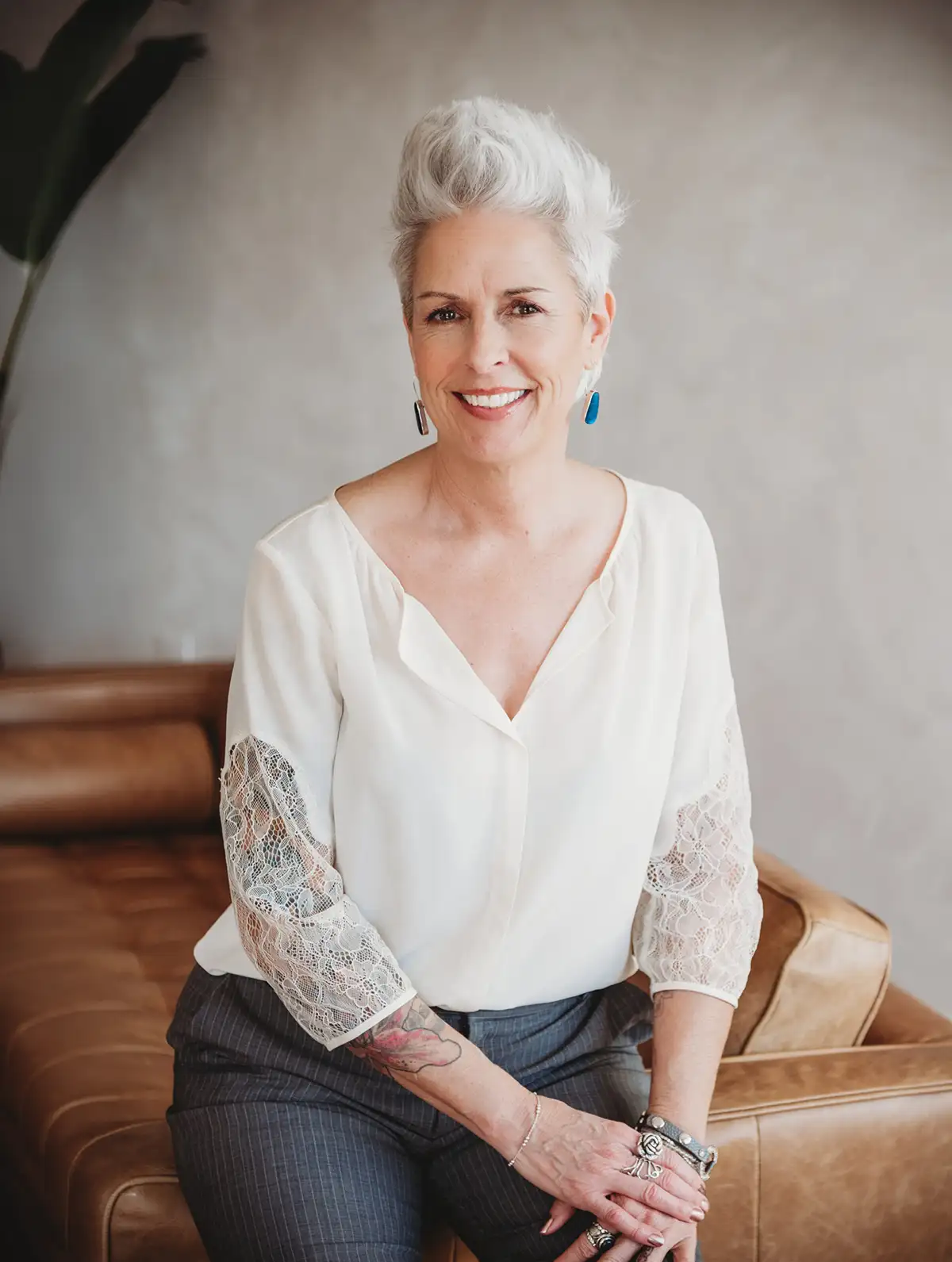 Nadine Hill, Dover NH bookkeeper and founder of Balance in Your Business, smiling warmly while seated on a rustic live-edge wooden bench, wearing a soft white cardigan and grey trousers, with short silver hair, sleeve tattoo visible, in a bright airy room with warm wood floors and a large plant in the background